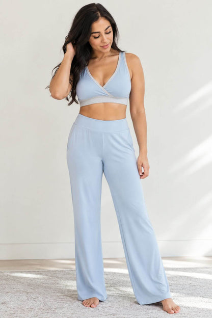 A woman modeling a light blue two-piece loungewear set consisting of a crossover sports bra and matching wide-leg pants, standing barefoot on a light rug against a white wall.