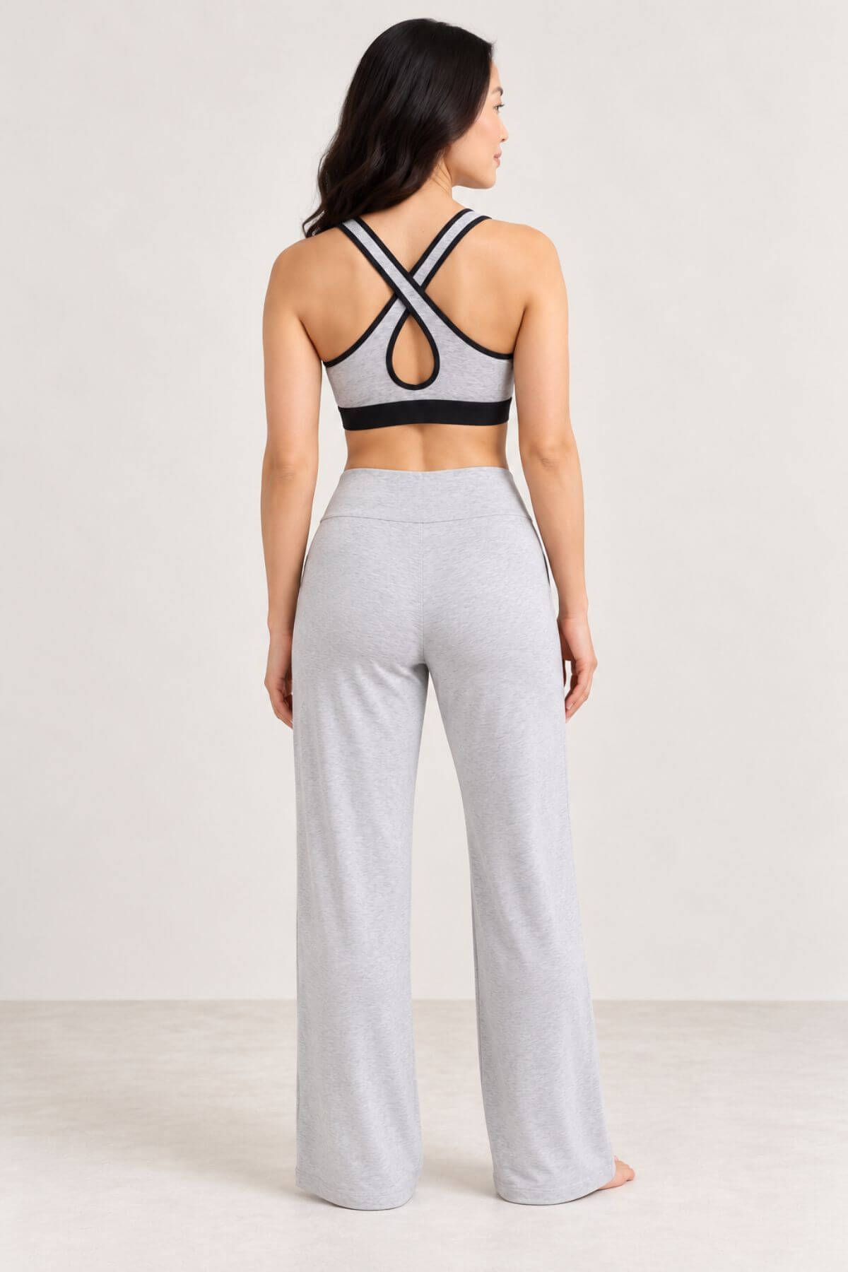 A woman shown from the back wearing a light gray and black sports bra and matching light gray wide-leg sweatpants in a studio setting.