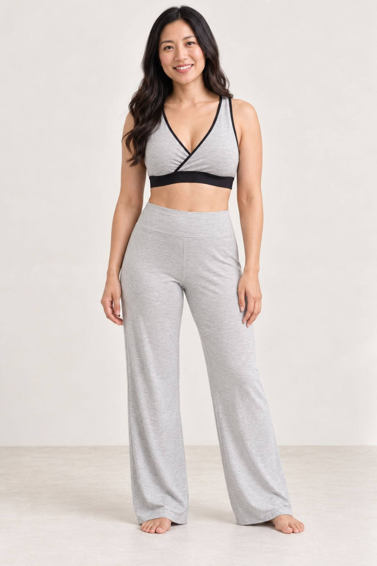 A woman modeling a light heather gray two-piece lounge set consisting of a wrap-style sports bra and matching high-waisted wide-leg pants, set against a plain light gray studio background.