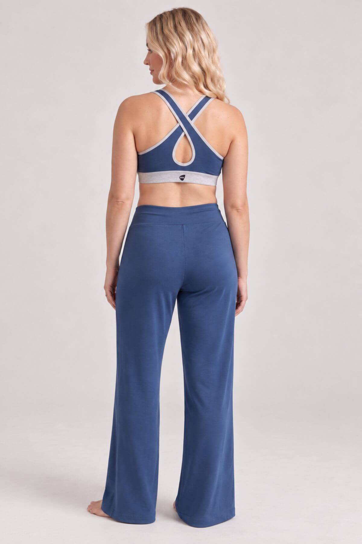 A woman shown from the back wearing a blue sports bra with a crossover crisscross back and matching blue wide-leg pants, photographed against a light gray studio background.
