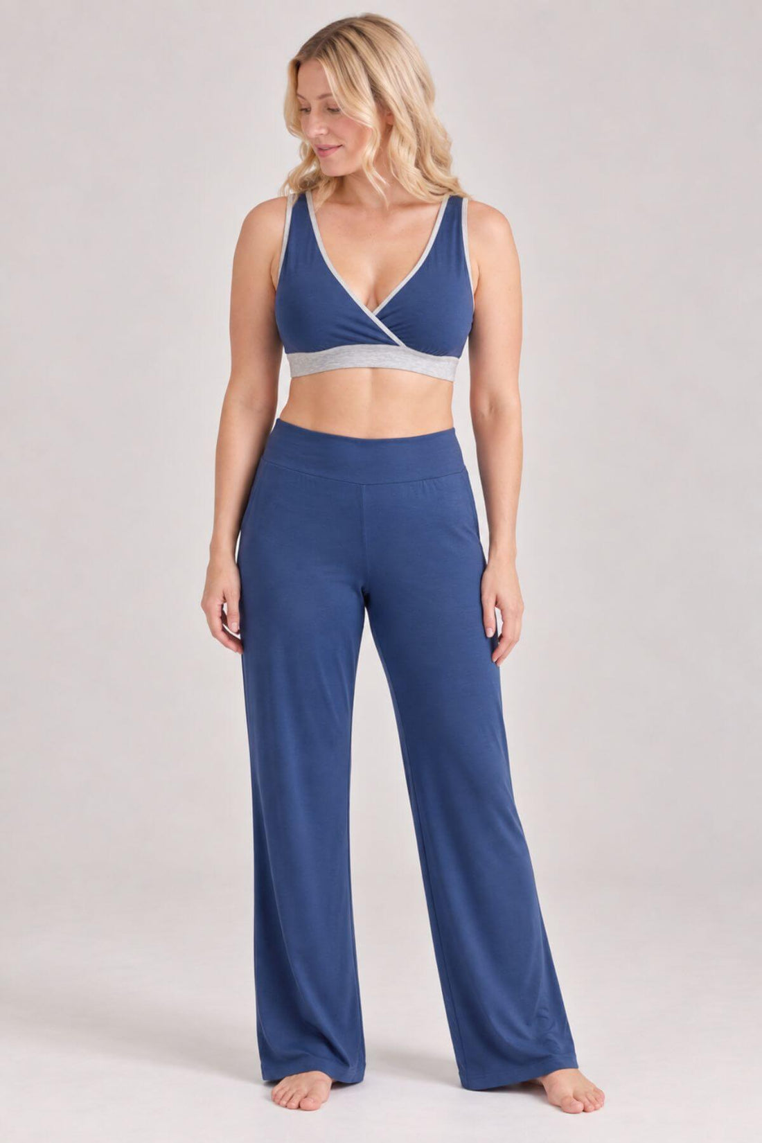 A woman modeling a matching two-piece loungewear set consisting of a blue wrap-style sports bra and high-waisted wide-leg pants, photographed against a neutral light gray studio 