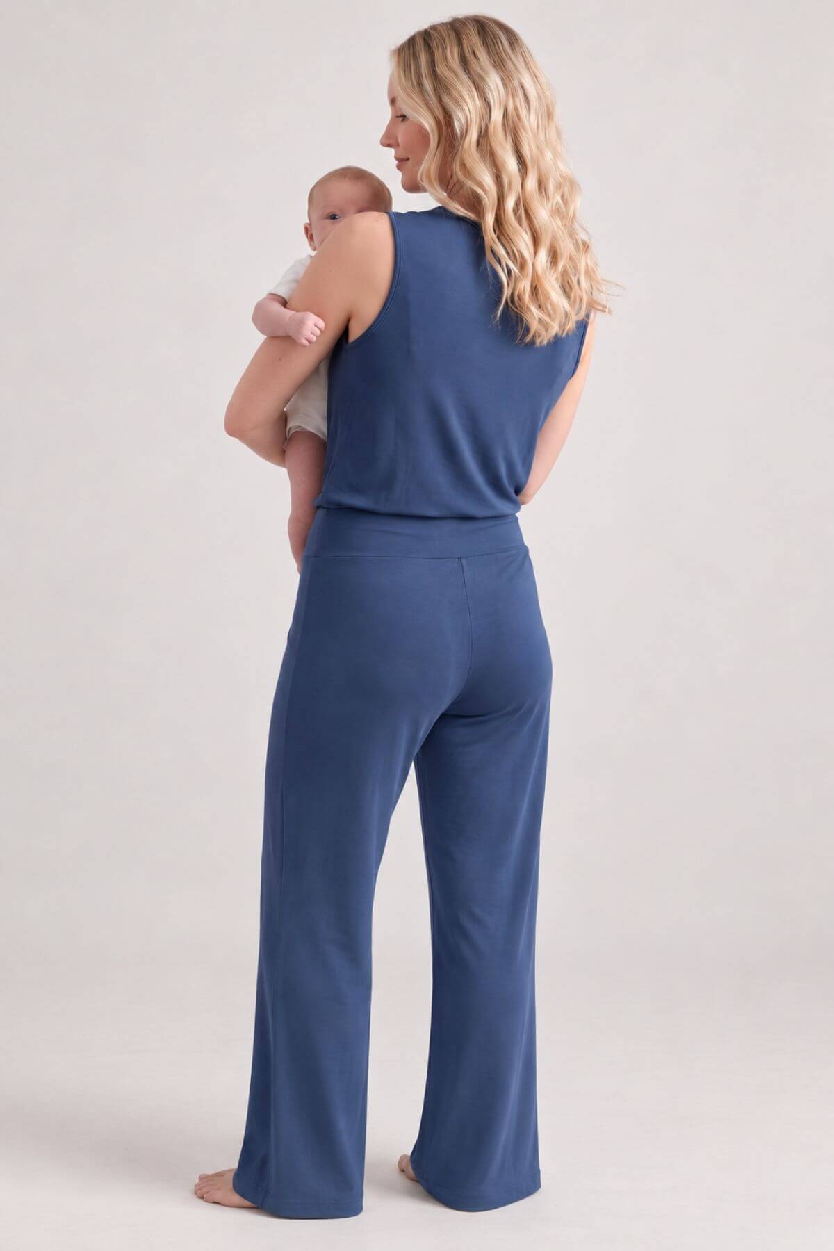 A woman wearing a sleeveless blue jumpsuit with wide legs, holding a baby, photographed from behind against a light gray studio background.