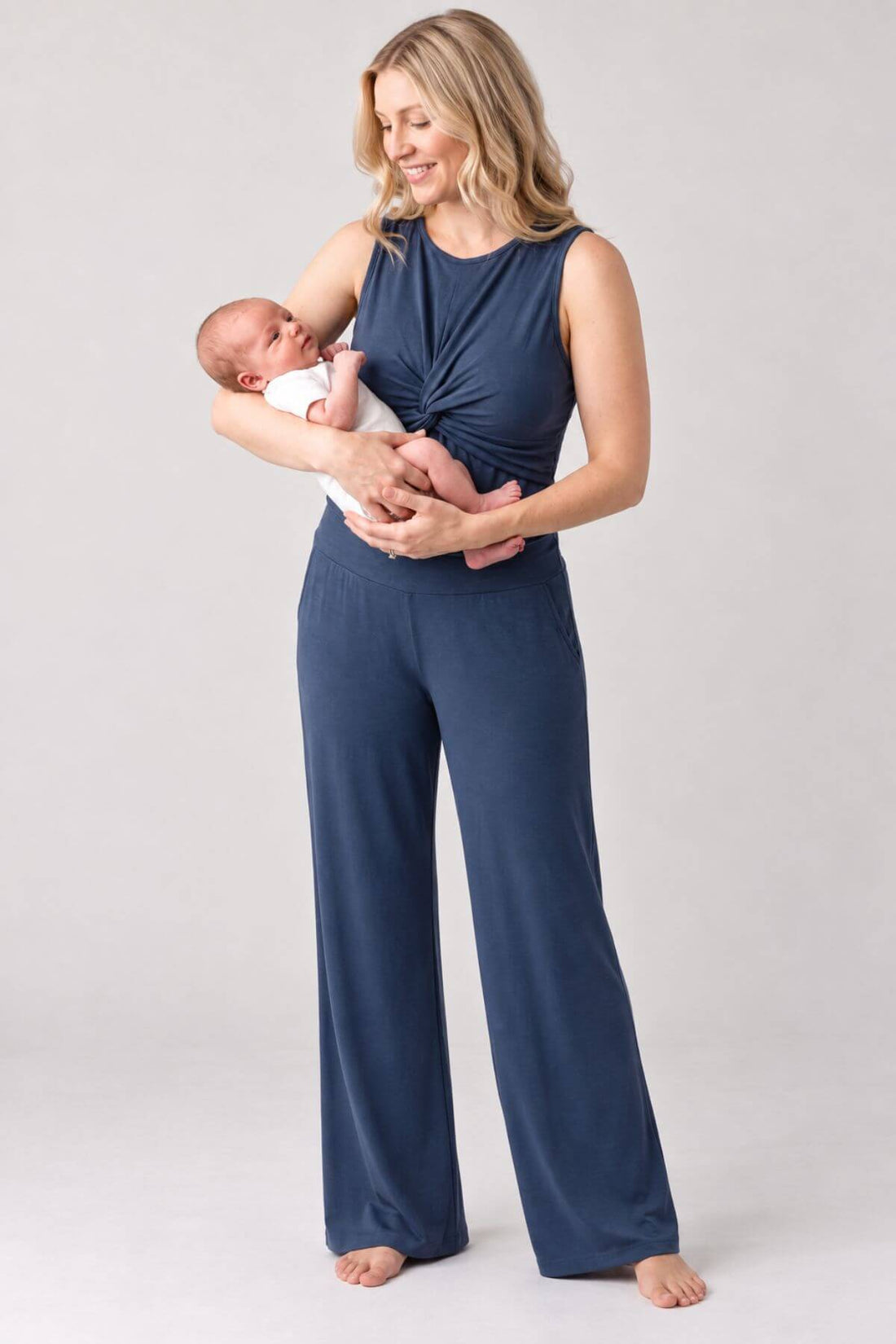 A woman holding a baby wearing a navy blue sleeveless jumpsuit with a twisted front detail and wide-leg pants, photographed against a light gray studio background.