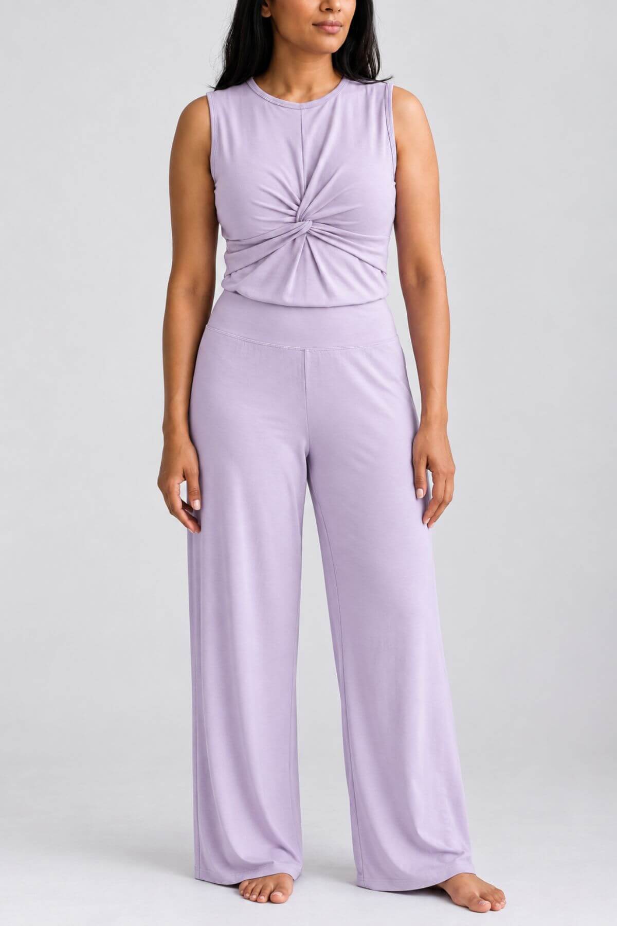 A woman modeling a sleeveless lavender jumpsuit with a gathered twist detail at the chest and wide-leg pants, photographed against a light gray studio background.