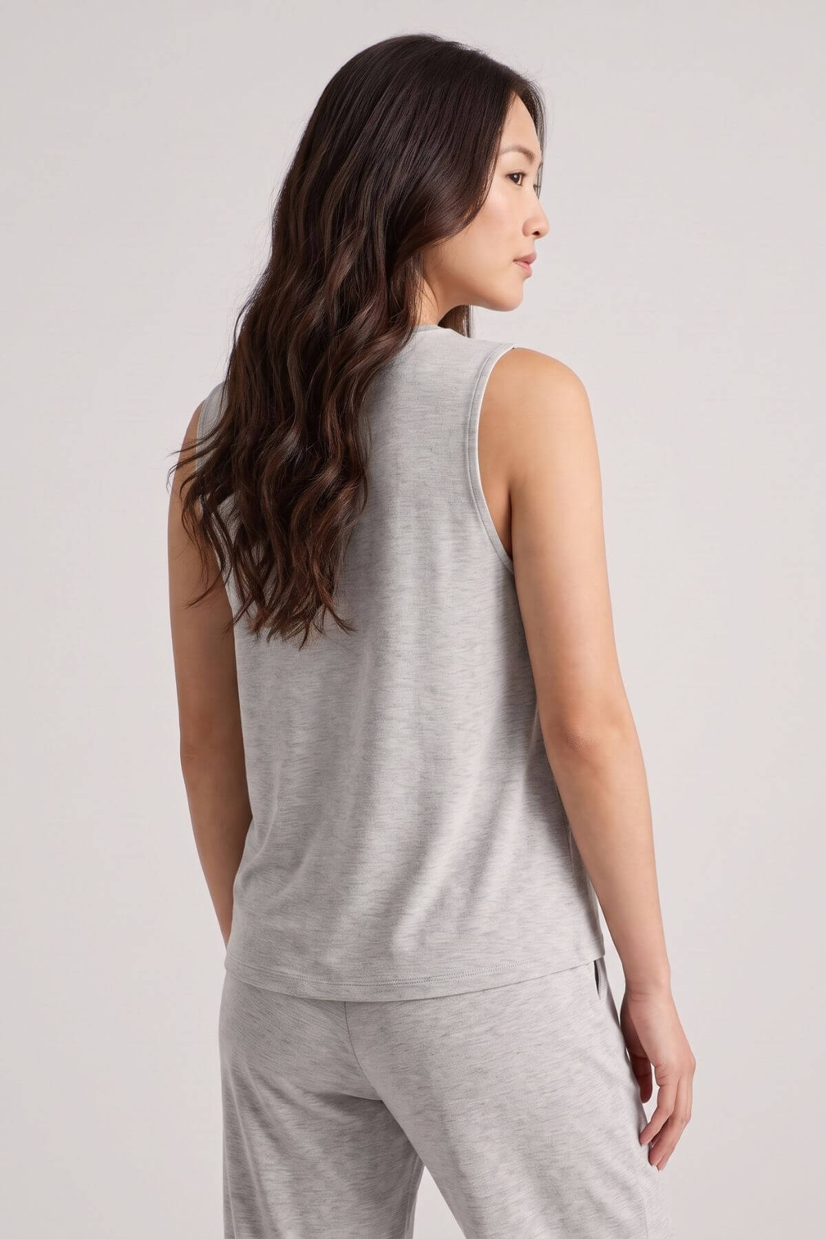 A woman with long dark wavy hair is shown from the back wearing a light heather gray sleeveless top and matching gray pants against a plain light gray studio background.