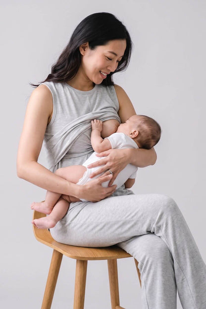 A woman sitting on a wooden stool breastfeeds a baby using a light gray wrap top and matching pants, photographed against a plain light gray studio background.