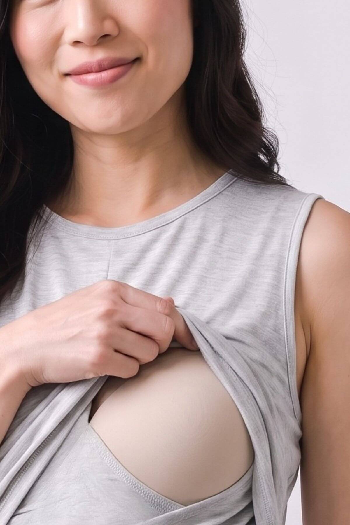 A close-up of a woman lifting the front of a light gray sleeveless top to reveal a beige underwire bra. She is smiling slightly, and the background is a clean, neutral light gray studio backdrop.