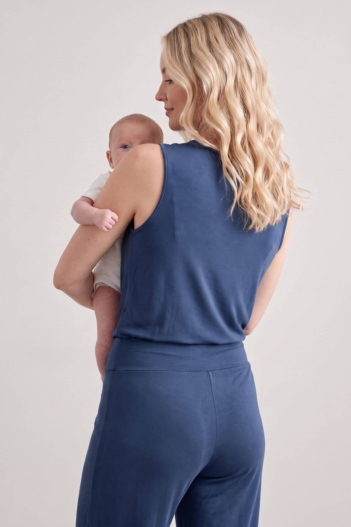 A woman with long blonde wavy hair, seen from behind, wearing a matching slate blue sleeveless top and high-waisted pants while holding a baby against her chest. The photo is taken in a studio setting with a plain light gray background.
