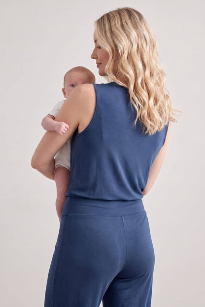 A woman with long blonde wavy hair, seen from behind, wearing a matching slate blue sleeveless top and high-waisted pants while holding a baby against her chest. The photo is taken in a studio setting with a plain light gray background.
