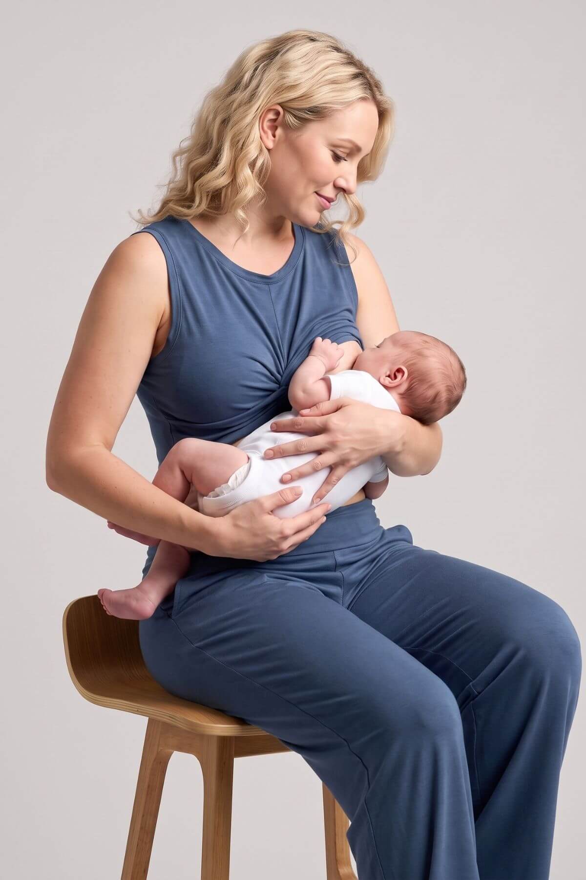 A woman in a navy blue nursing set sits on a wooden stool, breastfeeding a baby dressed in a white outfit against a neutral light gray background.