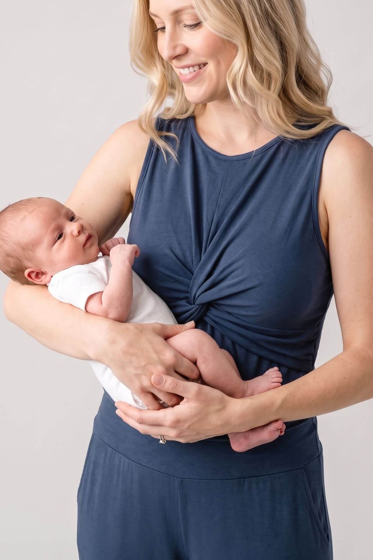 A woman in a navy blue sleeveless top and matching bottoms holds a sleeping newborn baby dressed in a white onesie against a plain light gray studio background.