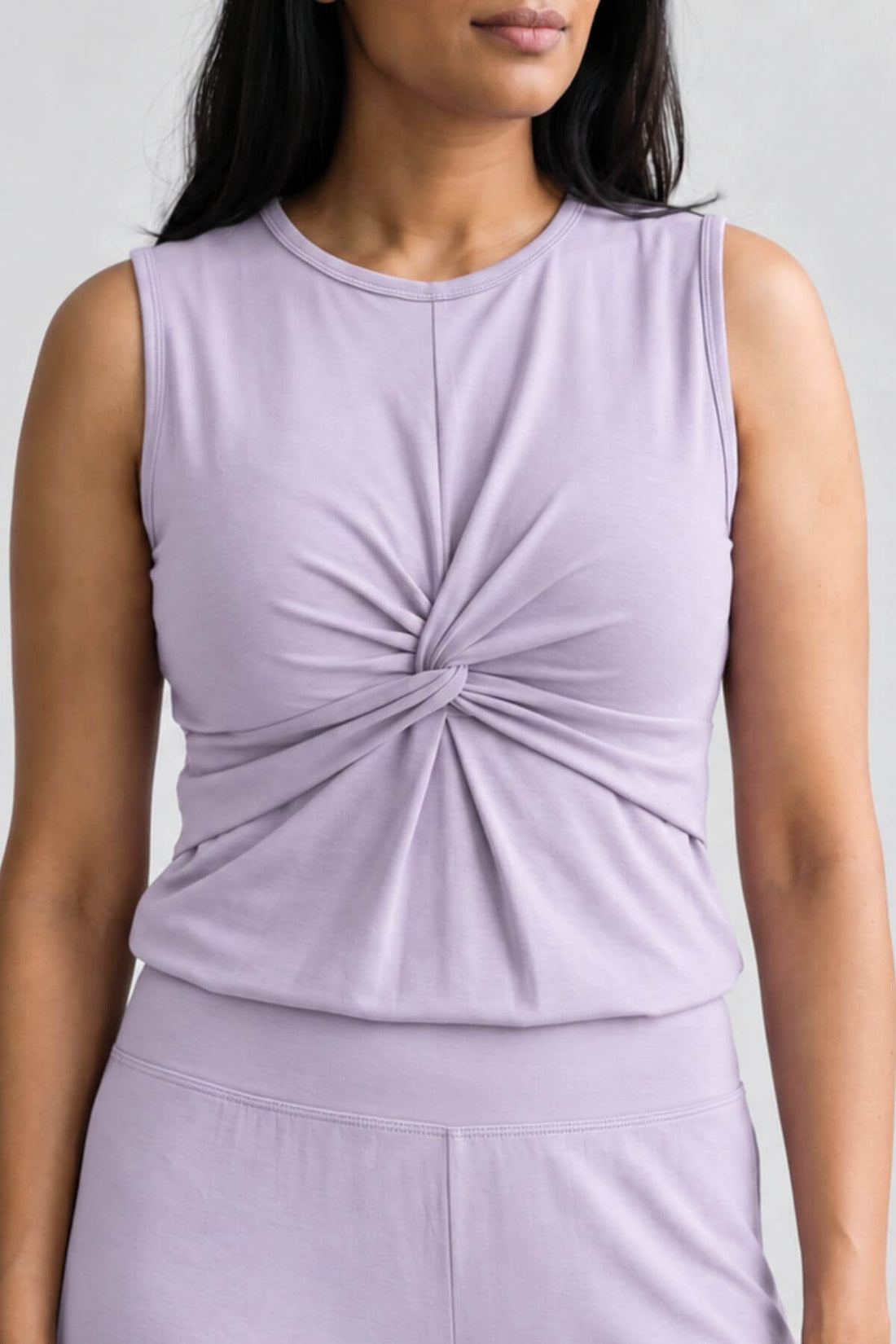 A close-up of a woman wearing a sleeveless light lavender top with a twisted knot detail at the center of the chest, paired with matching bottoms against a plain light gray background.