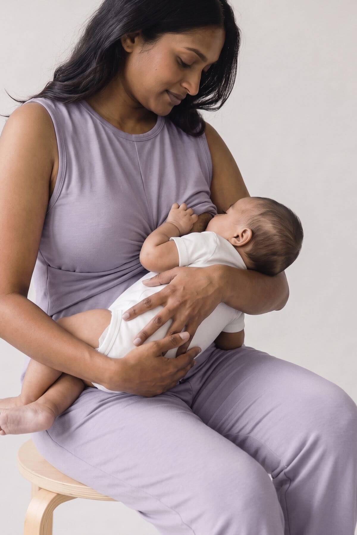 A woman in a lavender sleeveless top and matching pants sits on a wooden stool, breastfeeding a baby dressed in a white onesie against a plain light background.