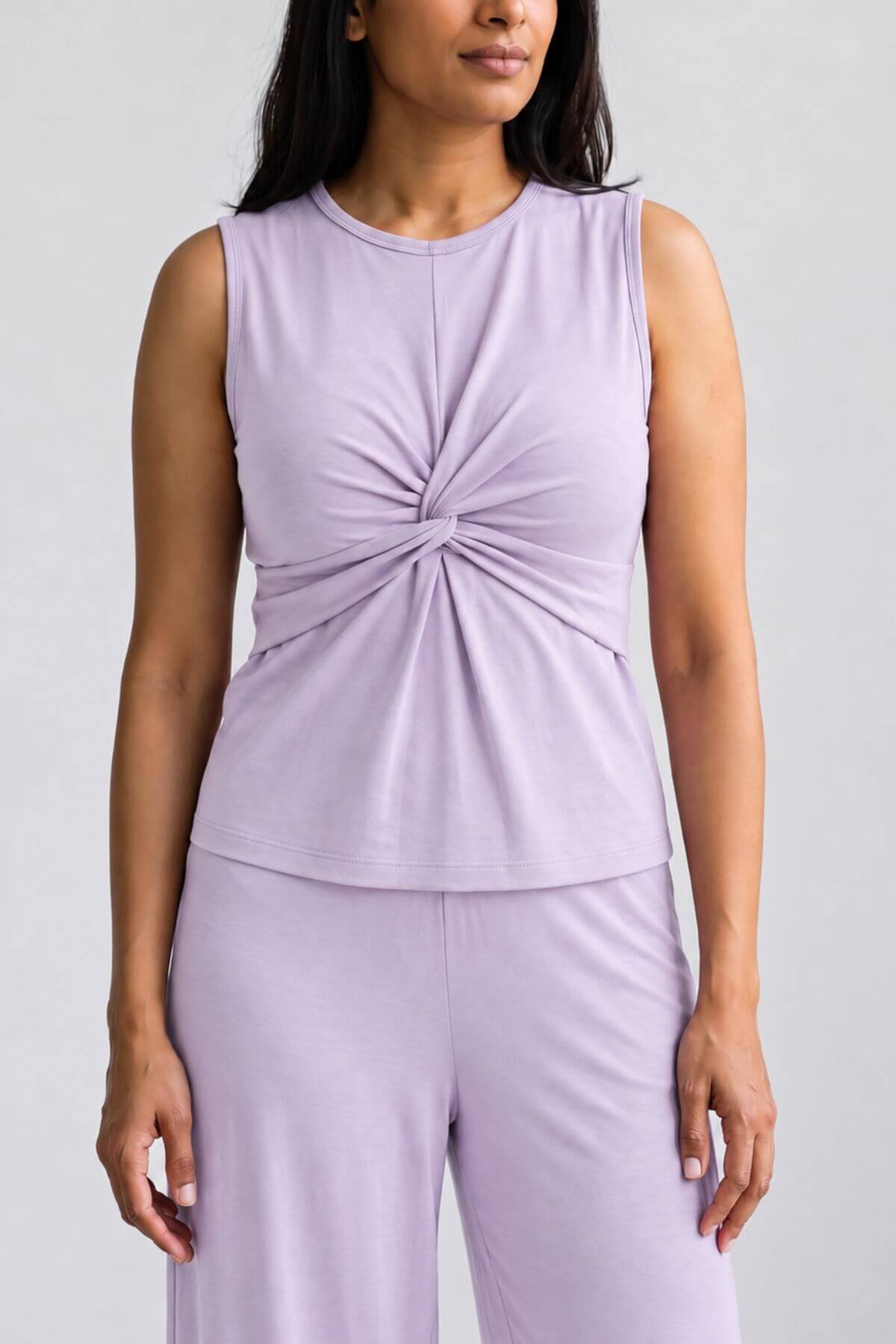 A woman wearing a sleeveless light purple top with a twisted knot detail at the center chest, paired with matching light purple pants, against a plain light gray background.