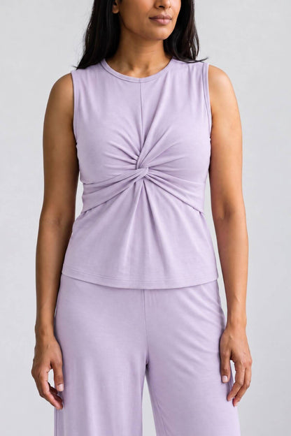 A woman wearing a sleeveless light purple top with a twisted knot detail at the center chest, paired with matching light purple pants, against a plain light gray background.