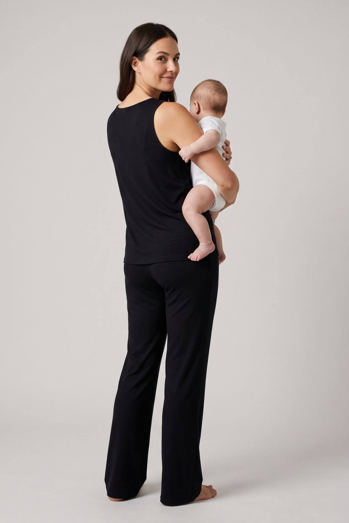 A woman wearing a black sleeveless top and black wide-leg pants holds a baby dressed in a white outfit, photographed against a plain light gray studio background.