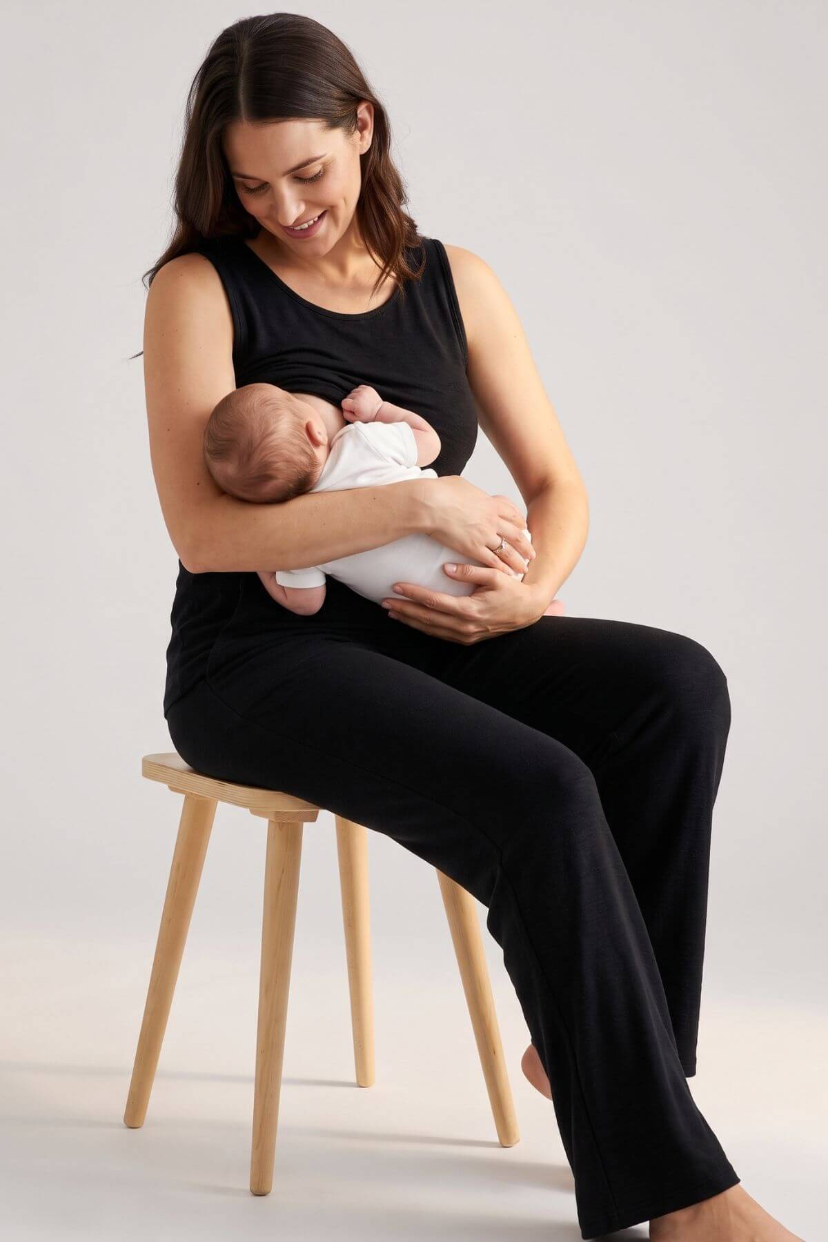 A woman in a black sleeveless top and black pants sits on a light wood stool, breastfeeding a baby wrapped in a white blanket, against a plain light gray studio background.