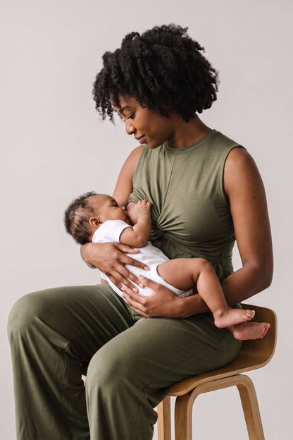 A woman in an olive green nursing top and matching pants sits on a wooden stool, holding a baby dressed in a white onesie against a plain light gray background.