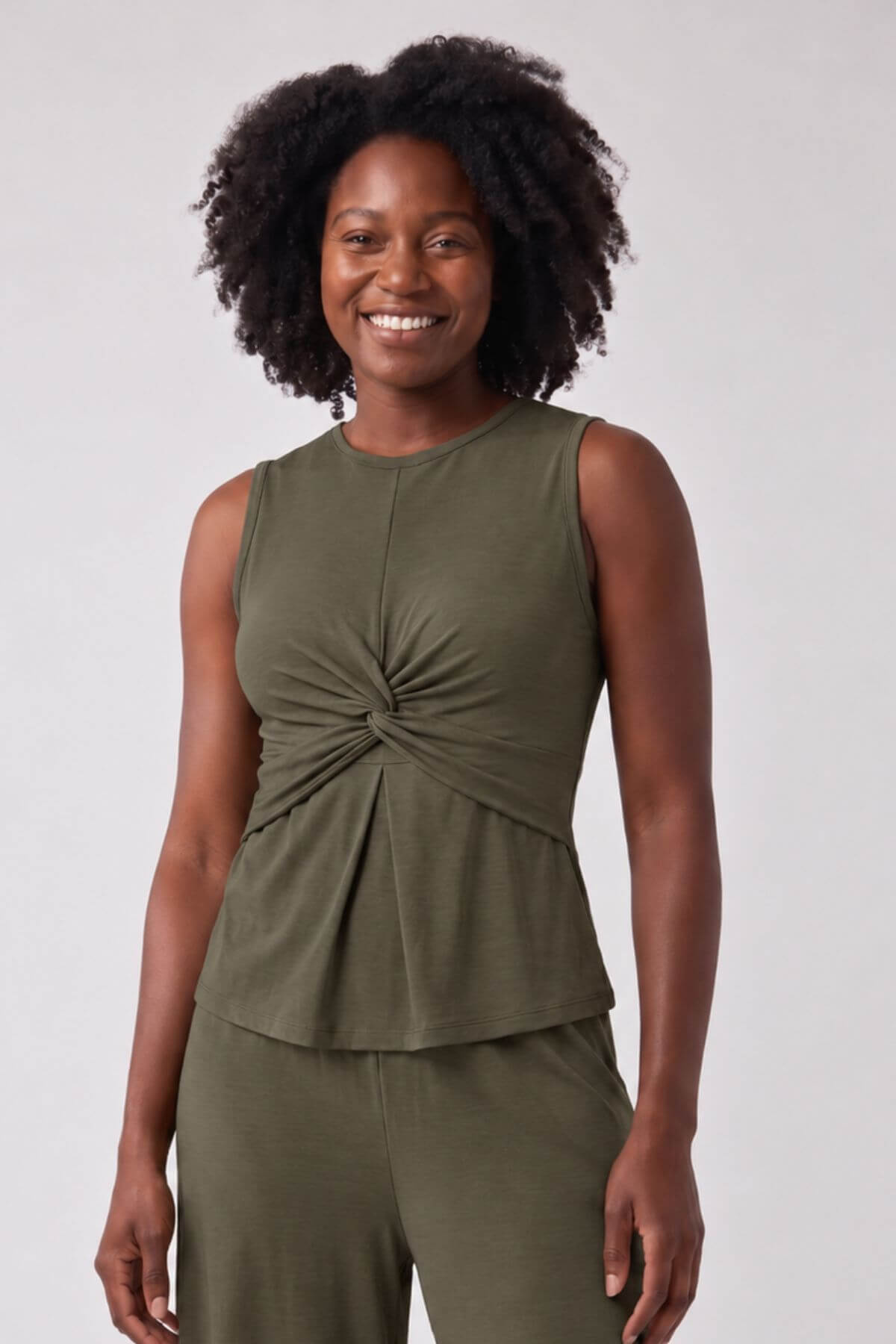 A smiling woman with curly black hair wearing an olive green sleeveless top with a twisted front detail and matching green pants, photographed against a light gray studio background.