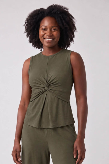 A smiling woman with curly black hair wearing an olive green sleeveless top with a twisted front detail and matching green pants, photographed against a light gray studio background.