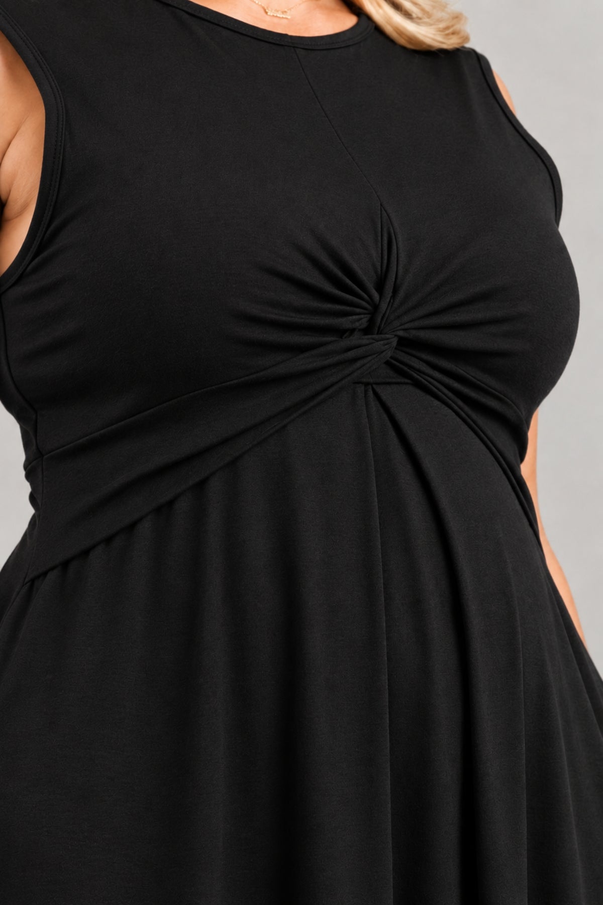 Close-up of a black sleeveless maternity dress with a twisted knot detail at the center bust, shown on a model against a light gray background.