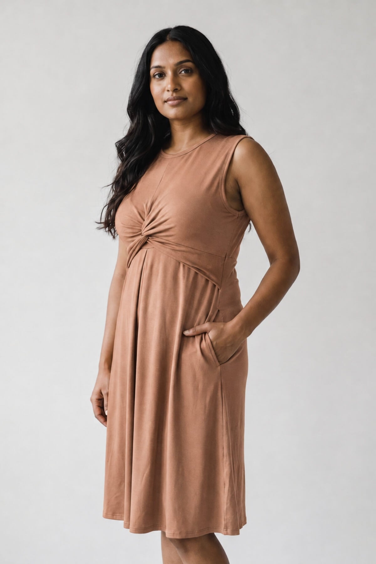 A woman modeling a sleeveless, knee-length dusty rose dress with a gathered twist detail at the chest and side pockets, photographed against a plain light gray studio background.