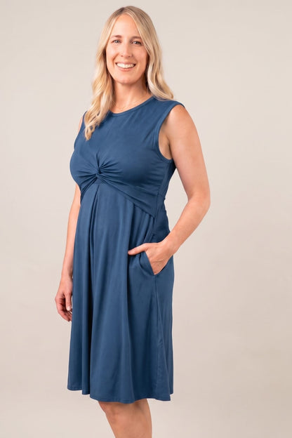A woman modeling a sleeveless blue A-line dress with a gathered twist detail at the bust and side pockets, photographed against a plain light beige studio background.