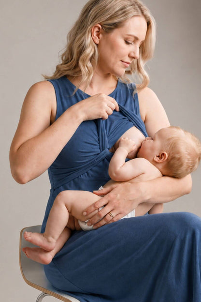 A woman wearing a blue nursing tank top and matching pants holds a baby while breastfeeding, seated on a chair against a neutral gray background.