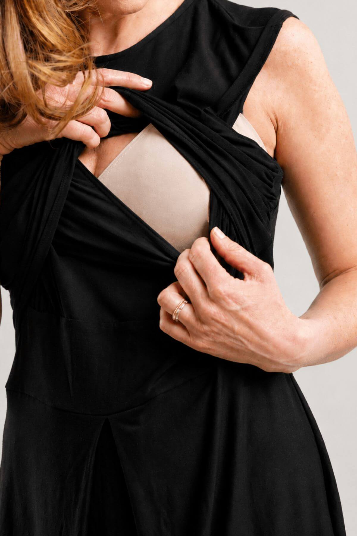 Close-up of a woman wearing a black maternity dress with a ruched, twist-front design that reveals a nude undergarment, set against a plain light gray background.