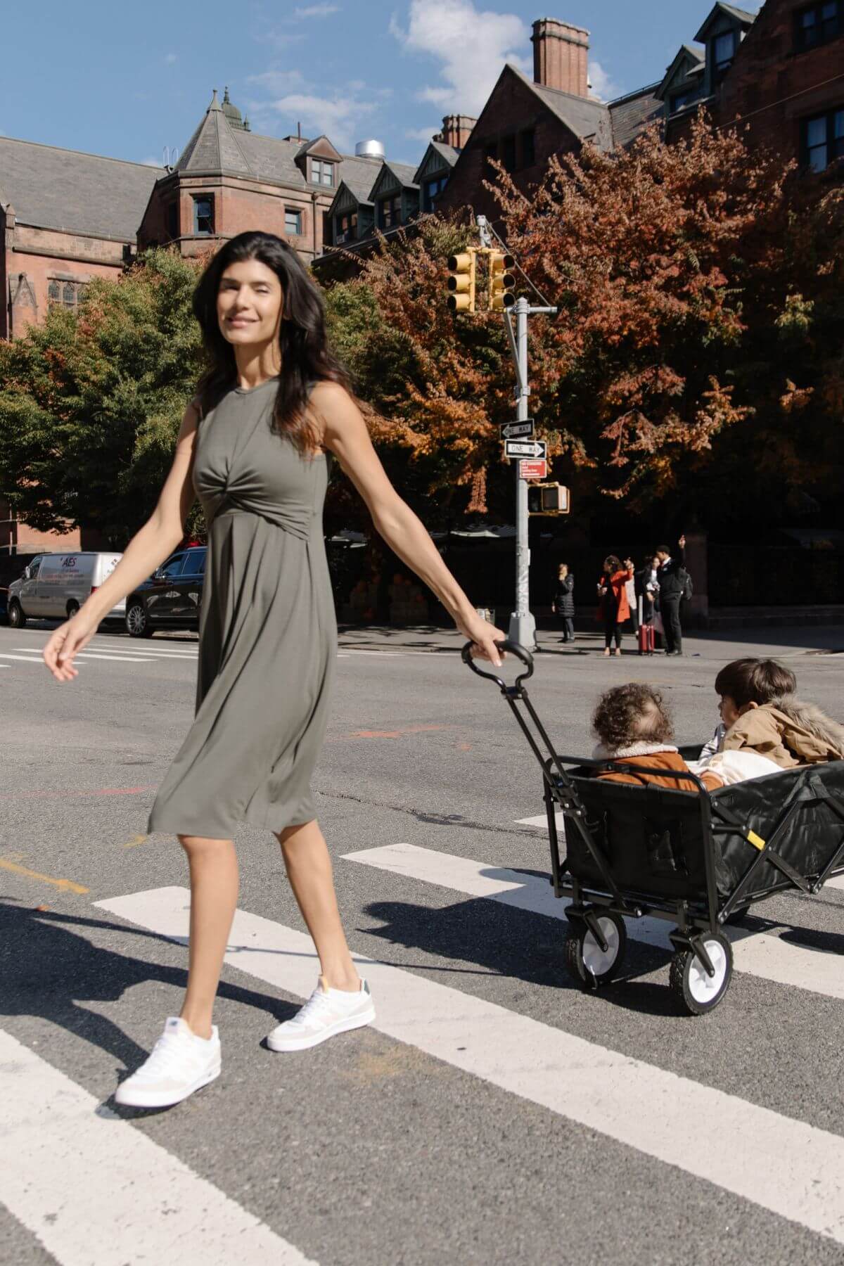 A woman in a sleeveless olive green midi dress walks across a city crosswalk while pulling a black wagon with two children in it. Brick buildings and autumn trees are visible in the background.