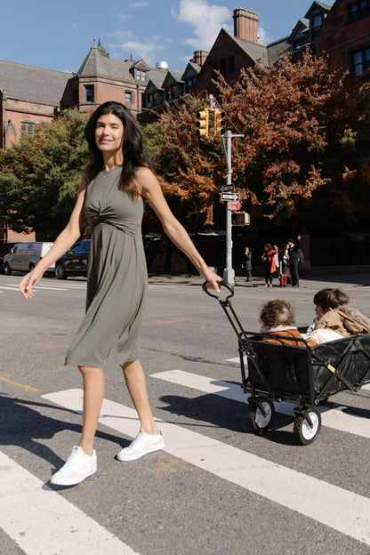 A woman in a sleeveless olive green midi dress walks across a city crosswalk while pulling a black wagon with two children in it. Brick buildings and autumn trees are visible in the background.