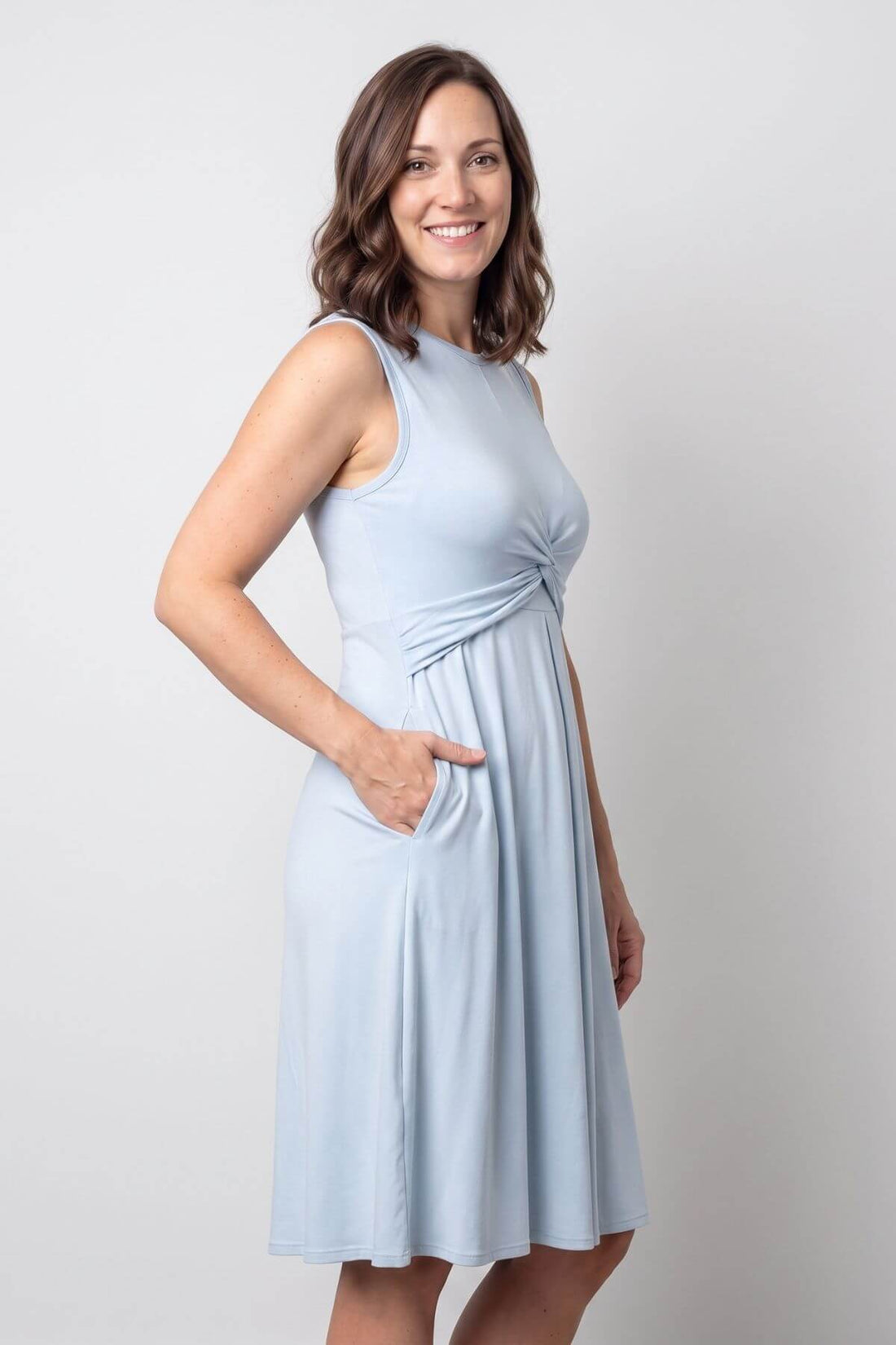 A woman modeling a sleeveless light blue A-line dress with a gathered twist detail at the waist and side pockets, photographed against a neutral light gray studio background.