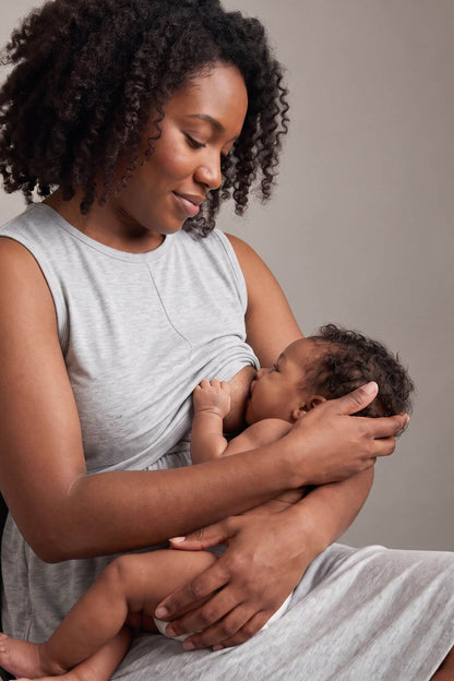 A woman with curly dark hair wearing a light gray sleeveless top and matching pants is breastfeeding a baby while seated against a plain taupe background.