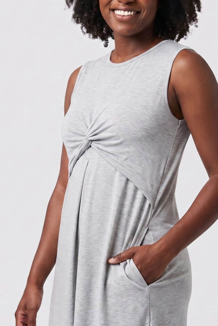 A woman with curly black hair wearing a light gray sleeveless knit dress with a twisted front detail and side pockets, posing against a light gray studio background.