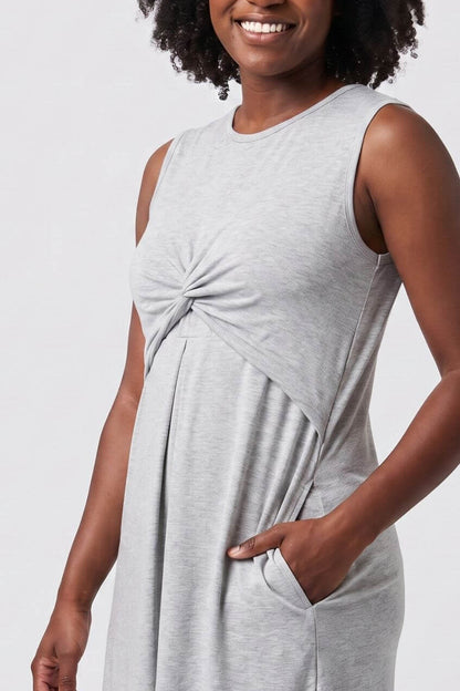 A woman with curly black hair wearing a light gray sleeveless knit dress with a twisted front detail and side pockets, posing against a light gray studio background.