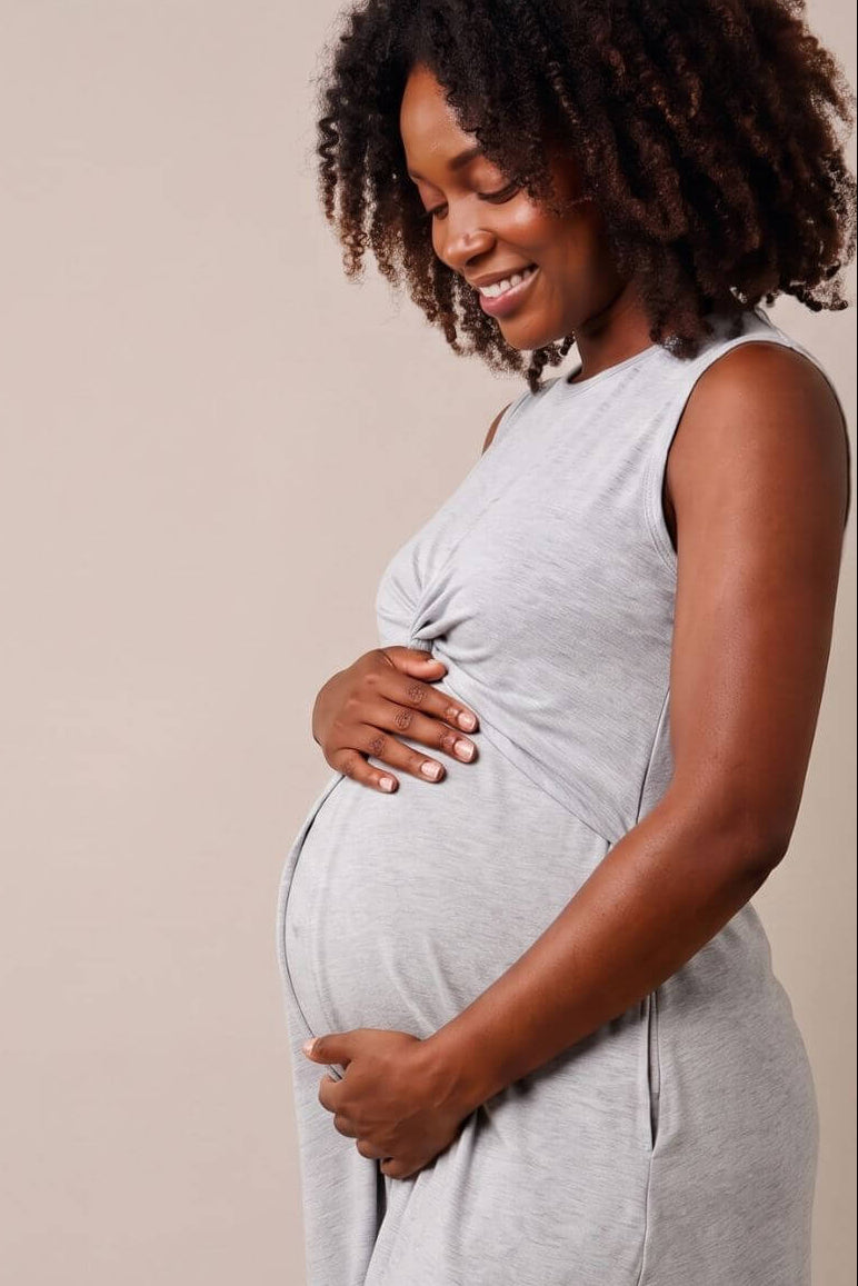 A pregnant woman with curly dark hair stands in profile wearing a light gray sleeveless maternity dress against a plain beige studio background.