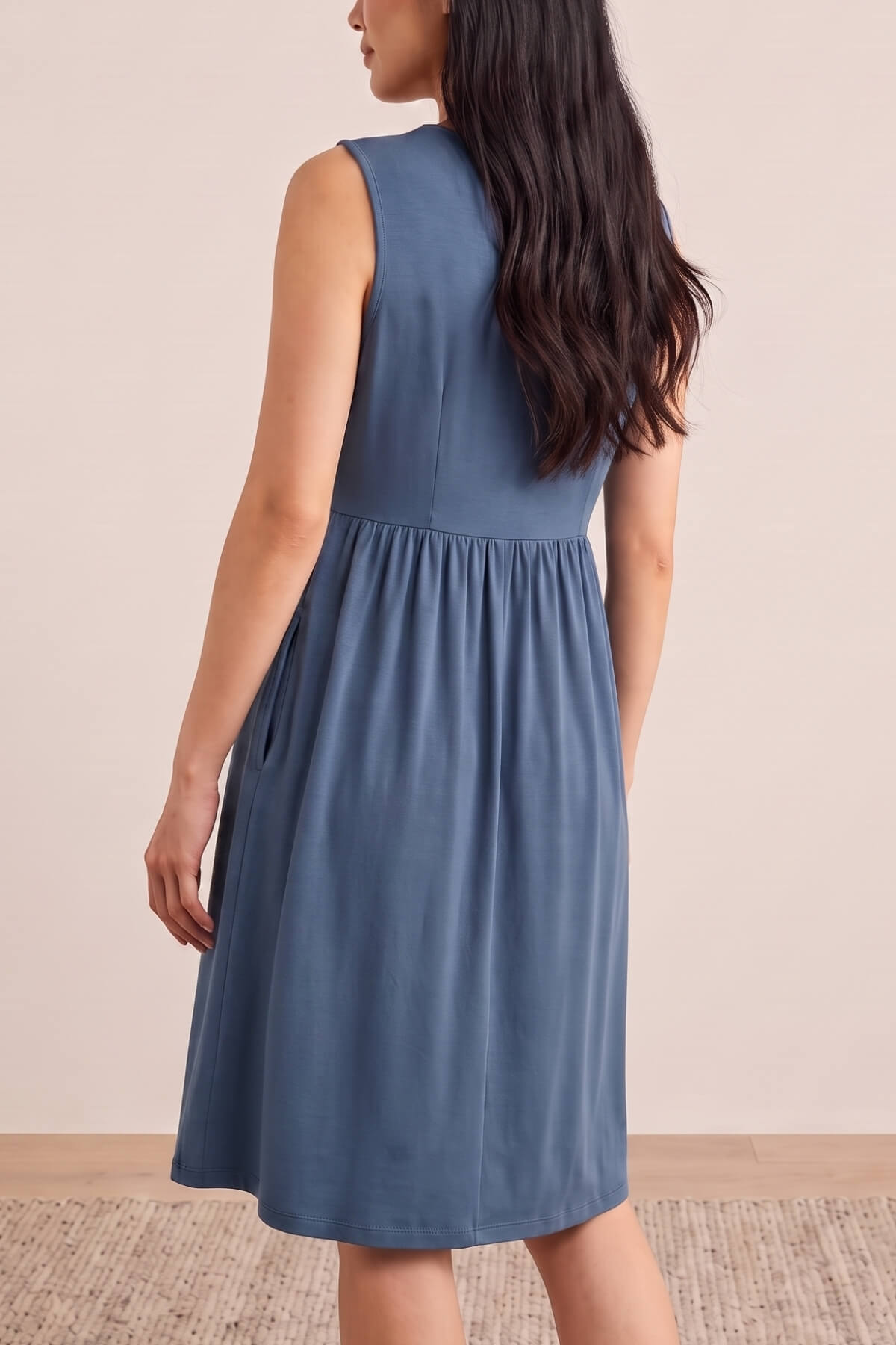 A woman wearing a sleeveless slate blue midi dress, shown from the back in a studio setting with a beige wall and light wood floor.