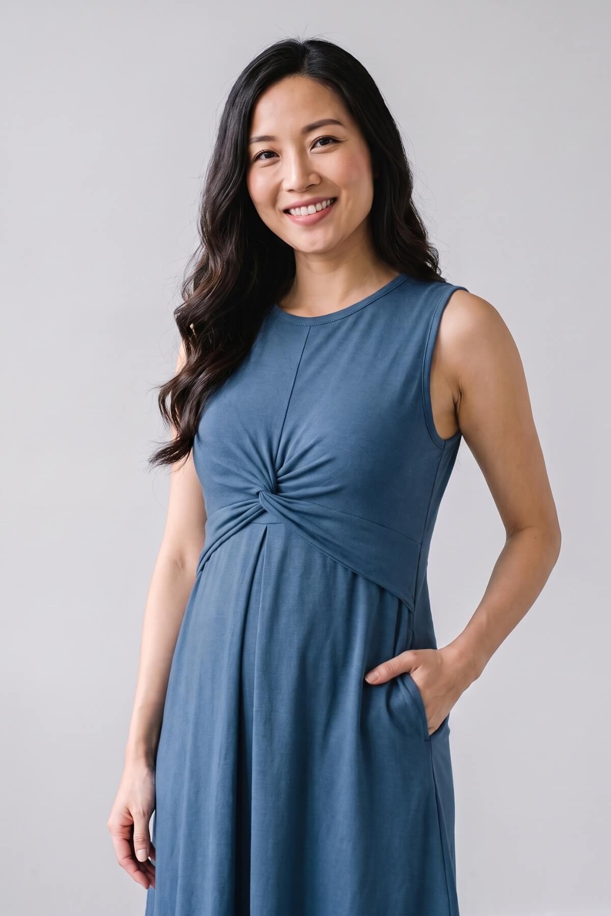 A woman wearing a sleeveless slate blue maternity dress with a twisted knot detail at the waist and side pockets, standing against a light gray studio background.