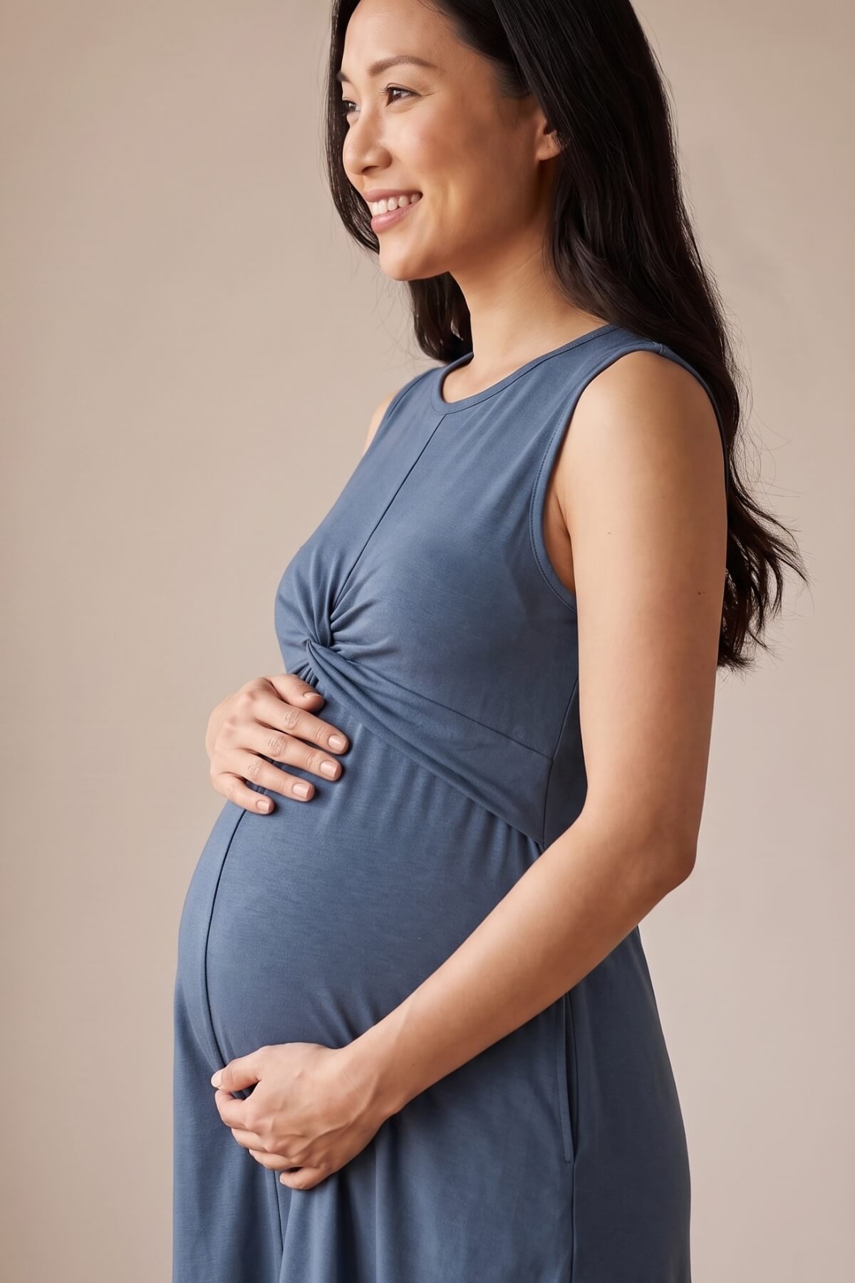 A pregnant woman wearing a sleeveless slate blue maternity dress, shown in profile against a solid beige studio background.