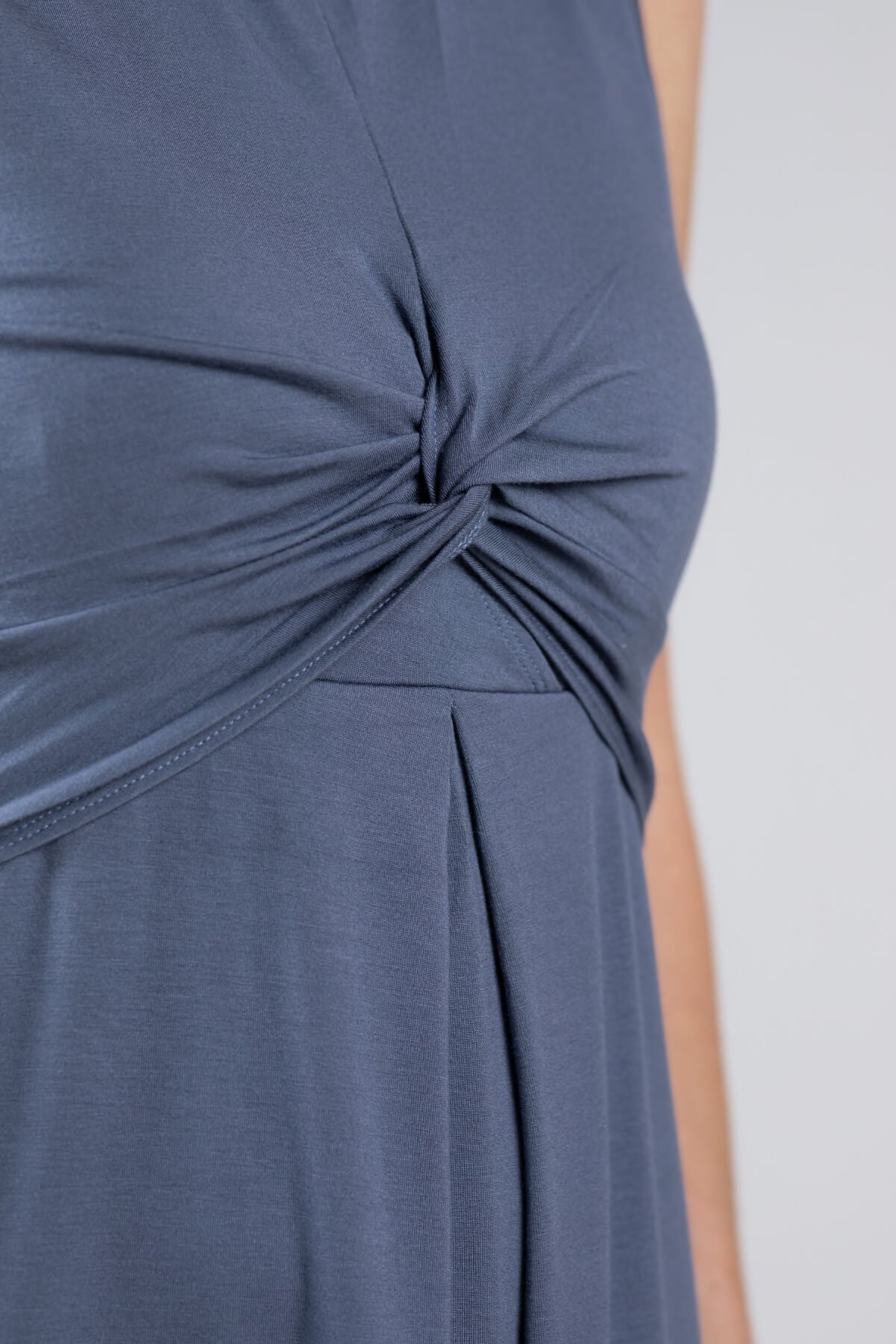 Close-up of a sleeveless dusty blue dress featuring a twisted knot detail at the waist and a draped neckline, shown on a model against a light gray background.