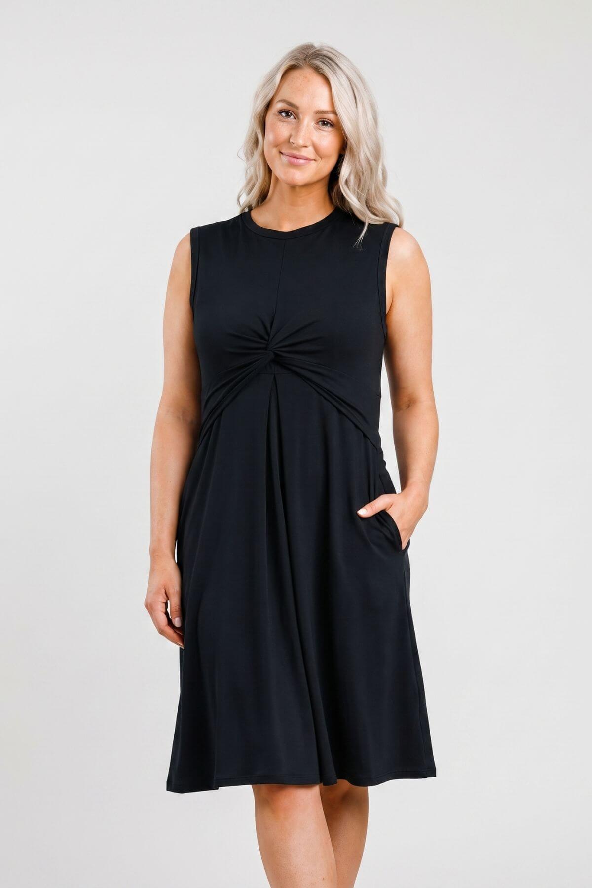 A woman modeling a sleeveless black A-line dress with a gathered twist detail at the center chest and side pockets, photographed against a light gray studio background..