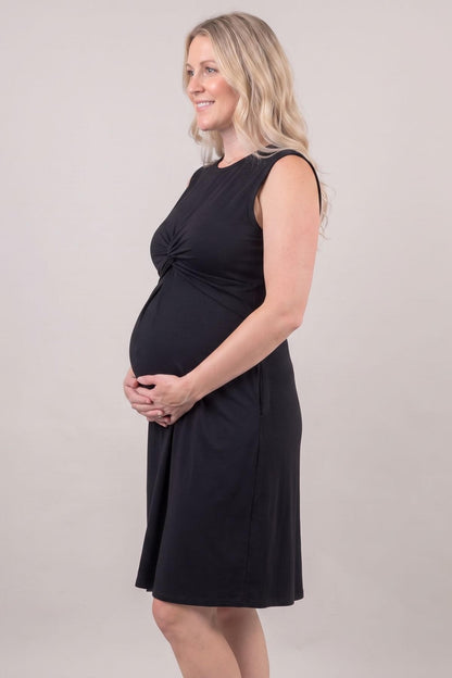 A pregnant woman with blonde hair is wearing a sleeveless black maternity dress with a gathered twist detail at the waist, standing against a neutral light gray studio background..