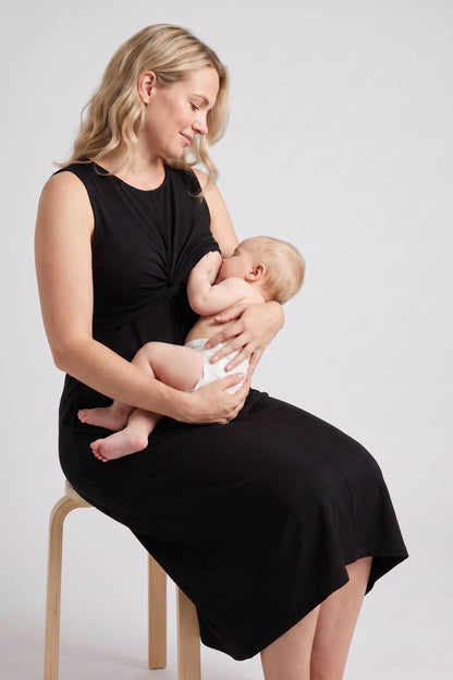 A woman in a black sleeveless dress sits on a wooden chair, breastfeeding a baby wearing a white diaper against a light gray studio background..