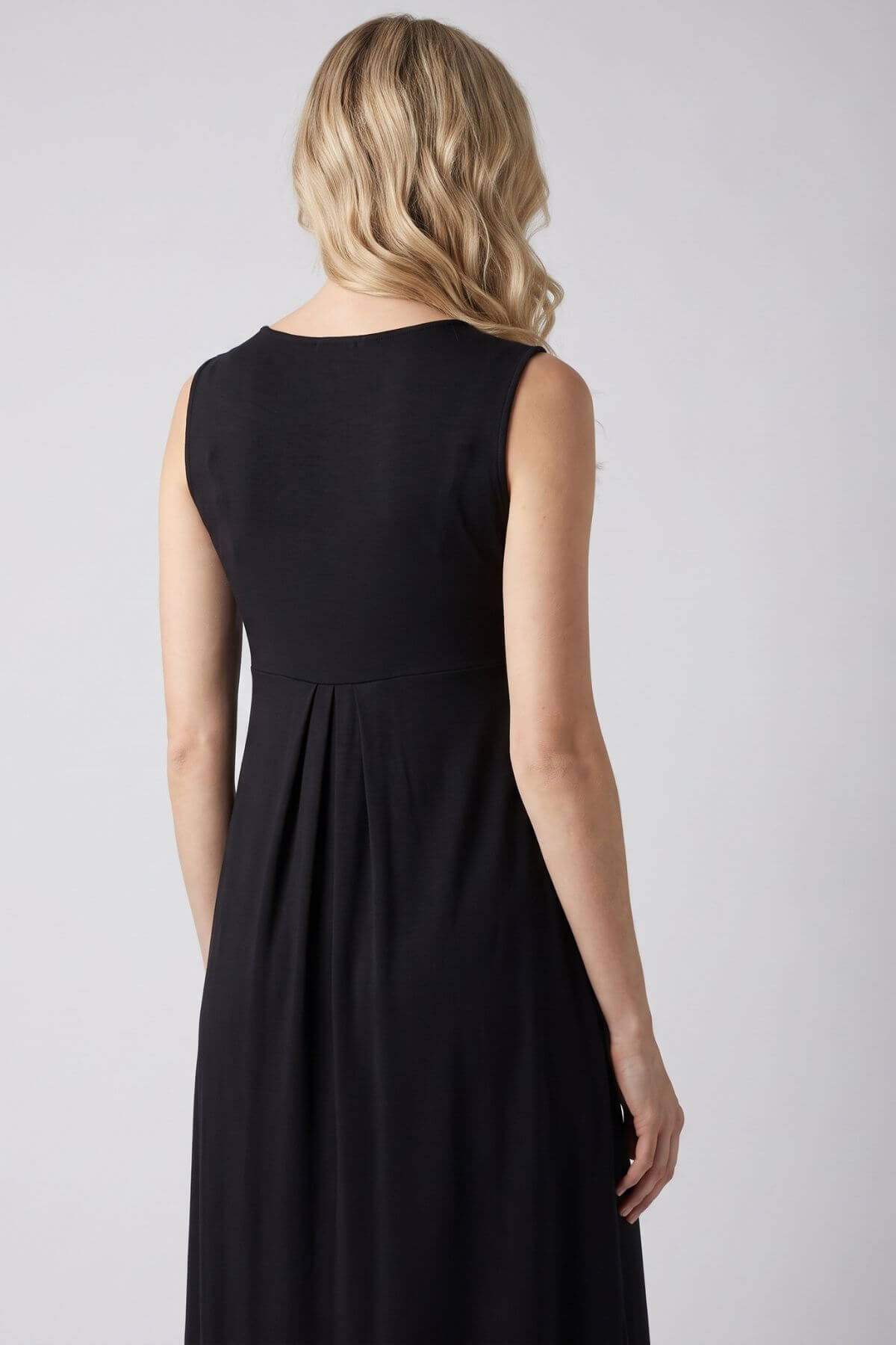 A woman with blonde hair is shown from the back wearing a sleeveless black midi dress against a light gray studio background..
