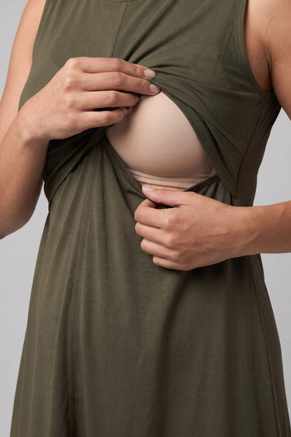 A close-up of a person wearing an olive green sleeveless dress and lifting the fabric to reveal a beige nursing bra underneath, set against a solid gray background.
