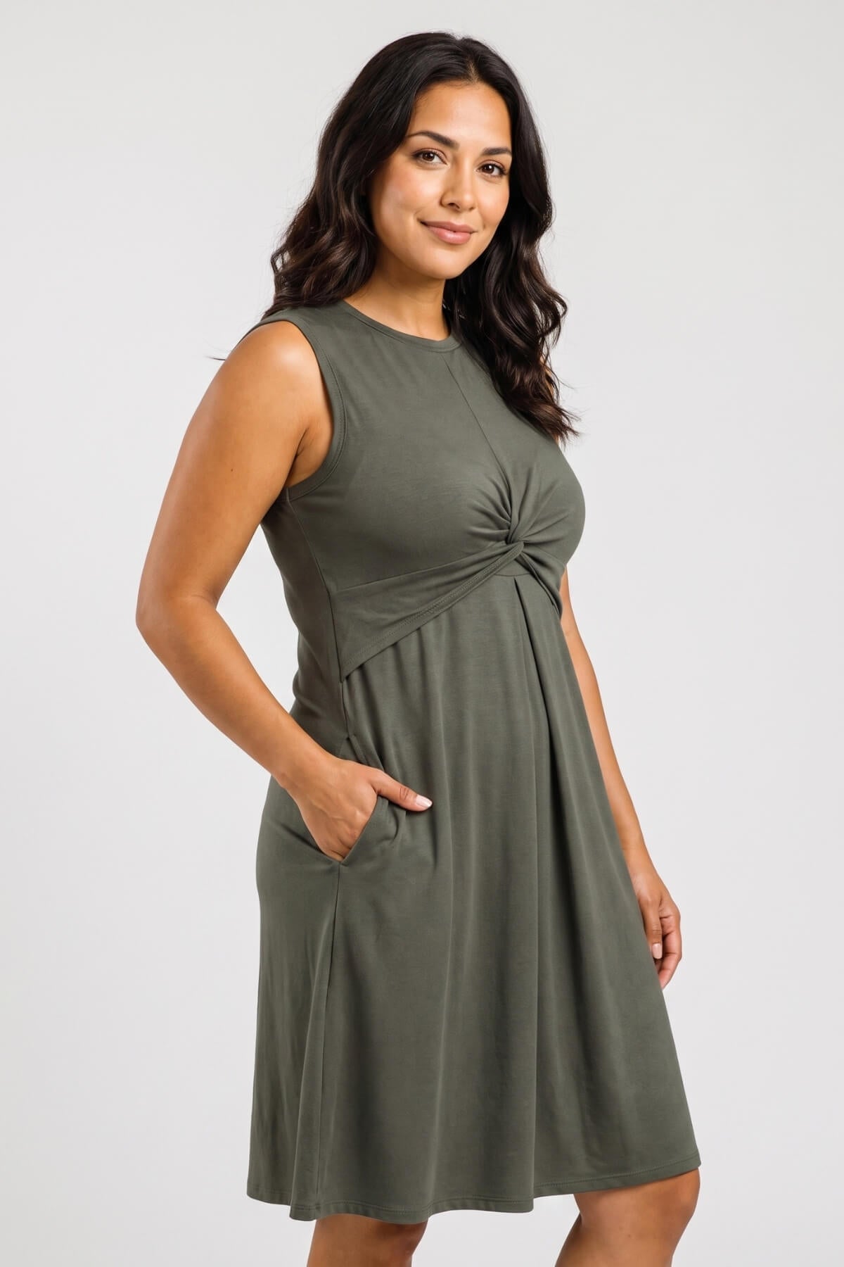A woman modeling a sleeveless olive green dress with a gathered twist detail at the waist and side pockets, photographed against a light gray studio background.
