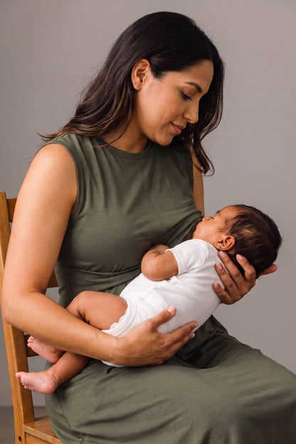 A woman in an olive green sleeveless dress holds and breastfeeds a newborn baby wearing a white onesie, seated on a wooden chair against a plain gray studio background.