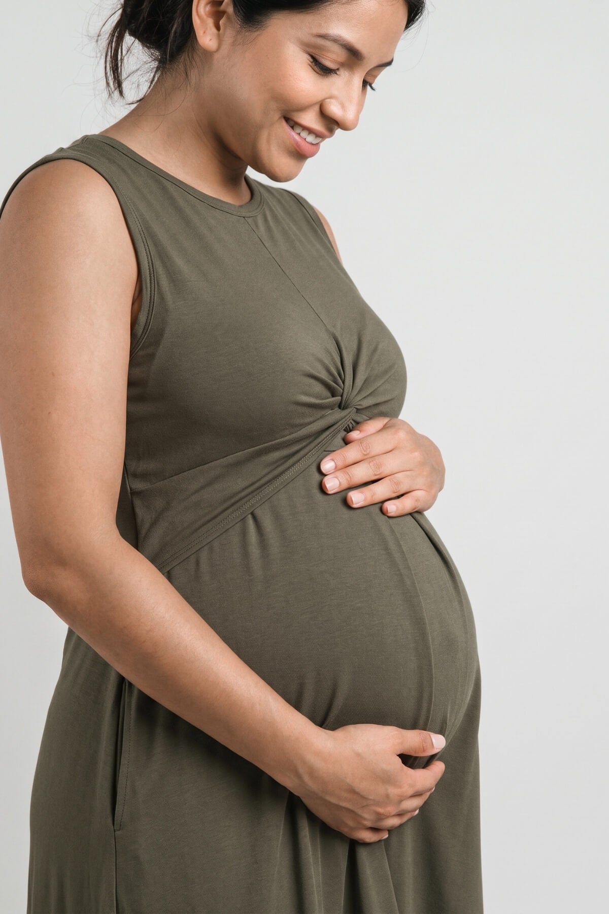 A pregnant woman wearing an olive green sleeveless maternity dress with a twisted detail at the bust, standing against a light gray background.