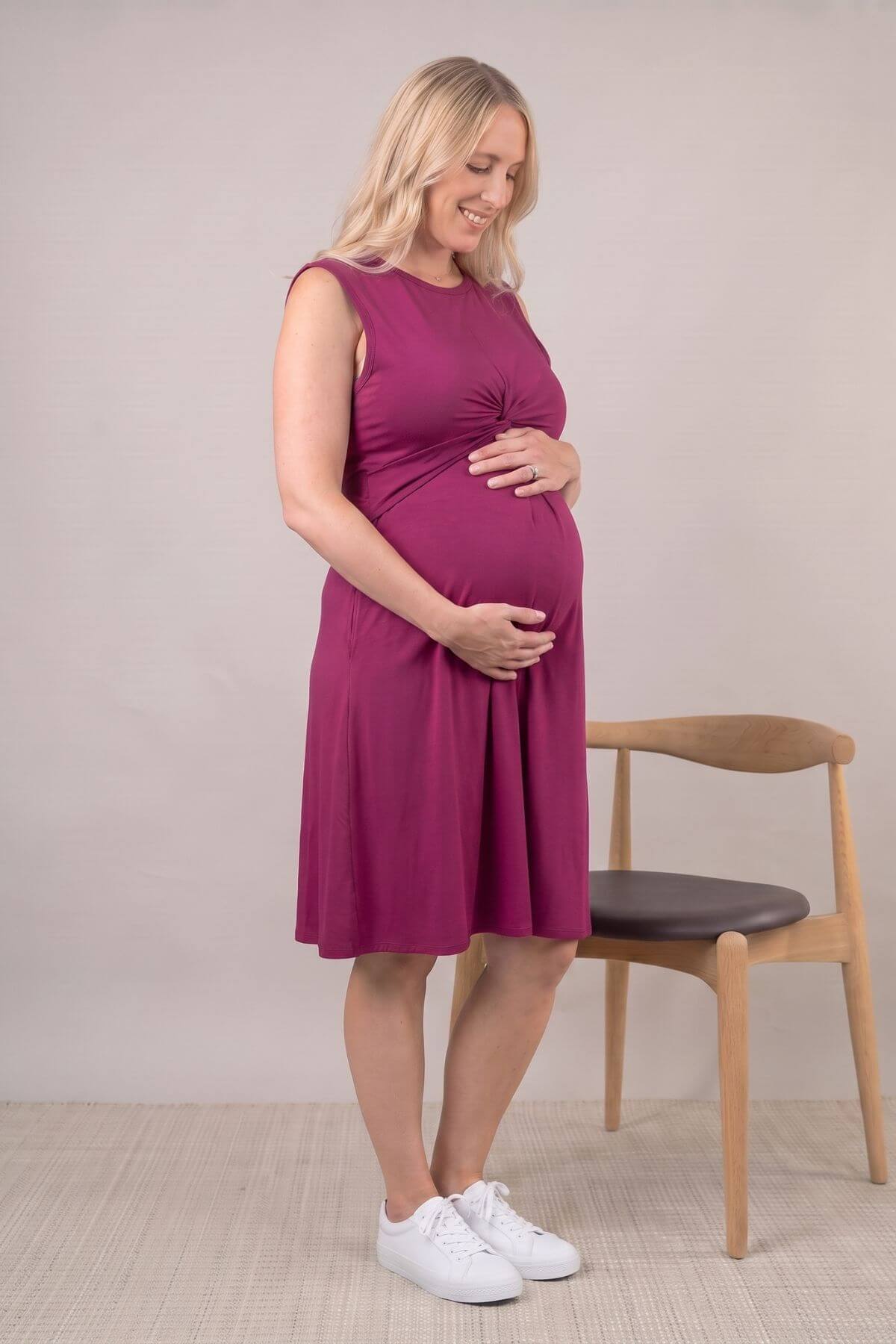 A pregnant woman wearing a sleeveless magenta maternity dress and white sneakers stands next to a wooden chair in a studio setting
