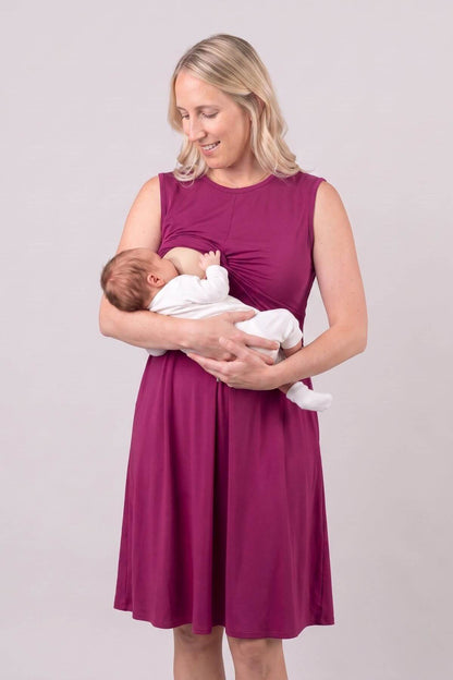 A woman wearing a sleeveless magenta maternity dress holds and breastfeeds a sleeping newborn baby dressed in a white outfit, set against a plain light gray studio background.